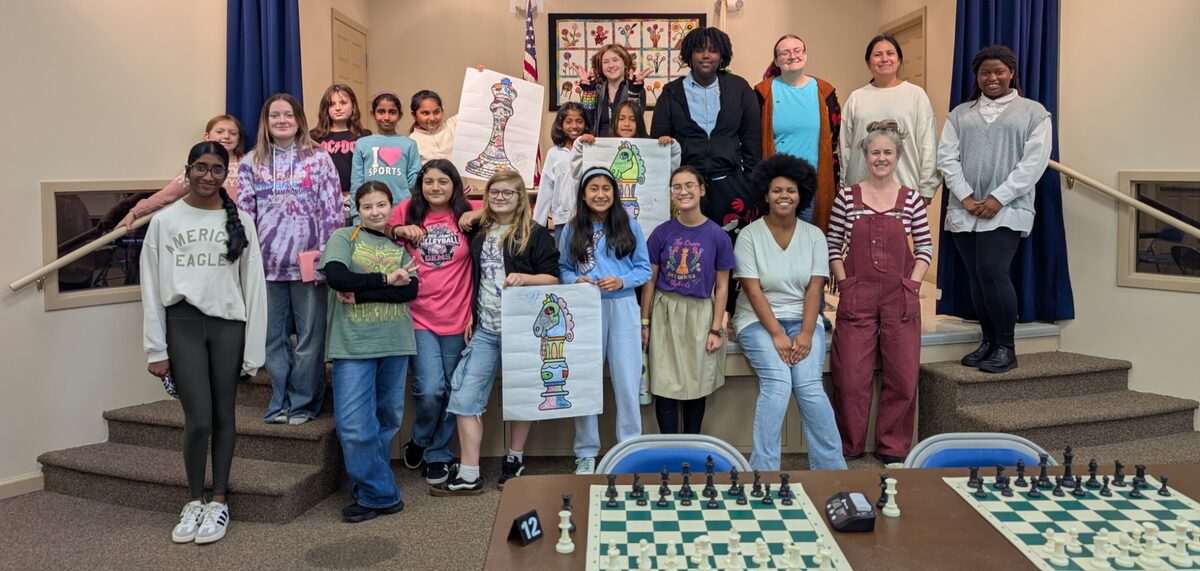 About twenty girls and women of all ages smiling together on a stage at the 2026 Kentucky Women and Girls Open, some holding posters of colored-in chess pieces, with chessboards set up in the foreground